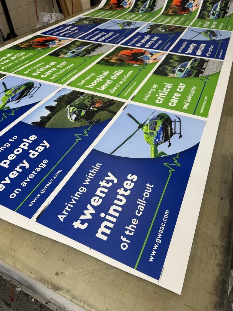 Full colour printed awareness posters for Great Western Air Ambulance featuring helicopters and emergency care messaging, laid out in a workshop.