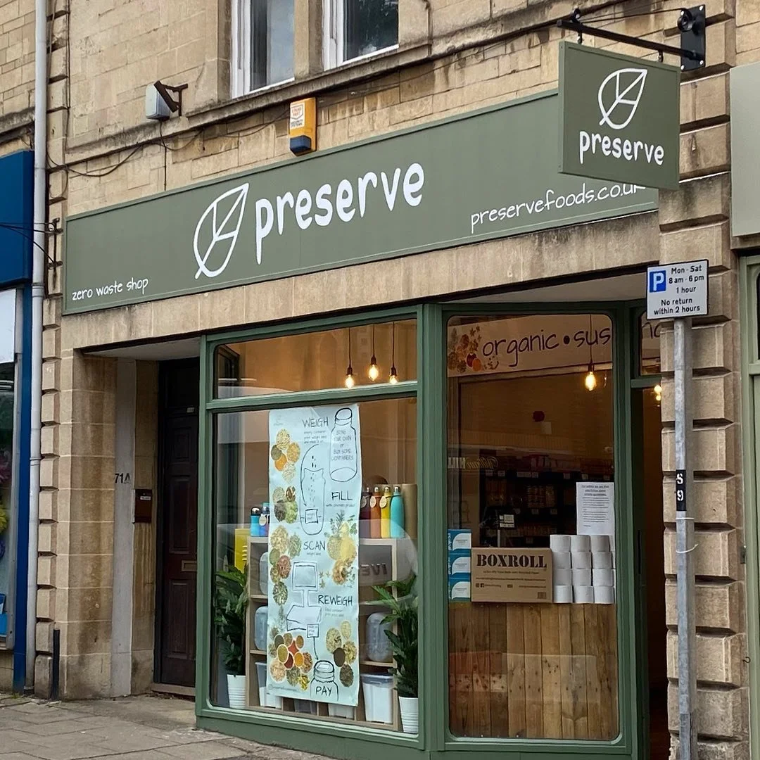 Storefront for Preserve zero waste shop with a green sign and window display featuring a poster with illustrations of food and instructions for how to use a scale, barcode scanner, and reweigh and pay options.