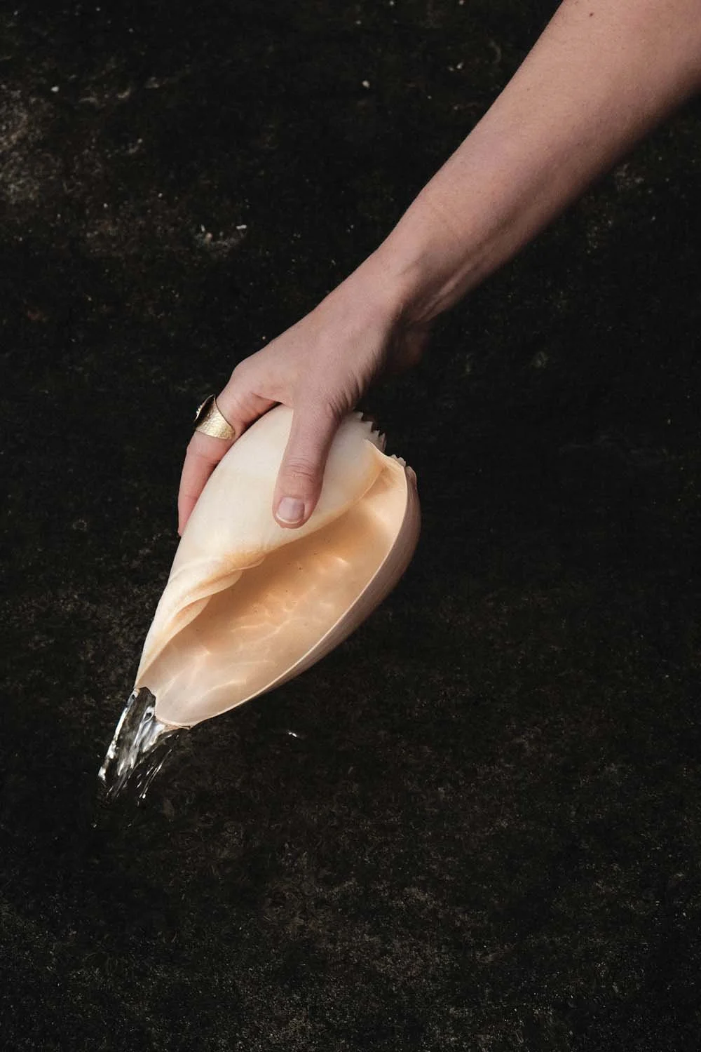 A person with a ring on their finger holding a seashell underwater, water flowing out from the shell onto a dark surface.