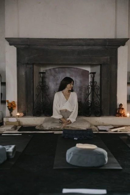 A woman sitting cross-legged in a meditation pose in front of a fireplace, surrounded by candles, books, and a Buddha statue.