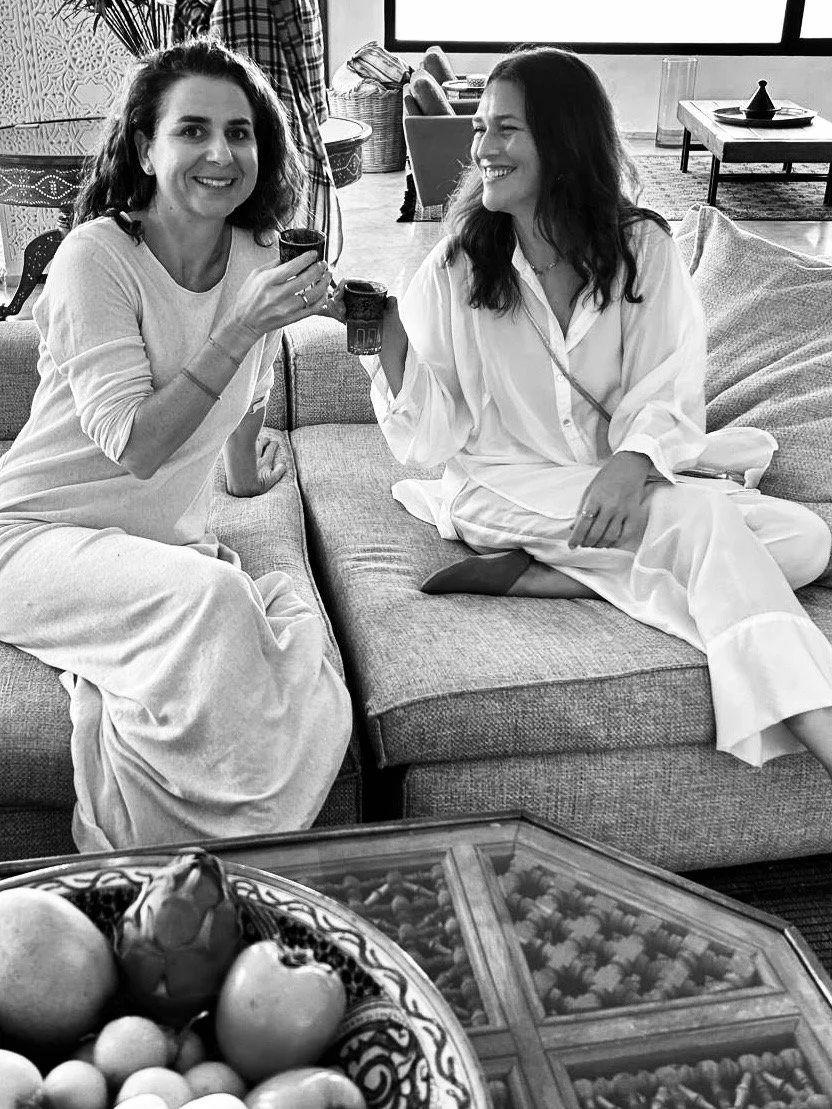 Two women sitting on a sofa, smiling and holding drinks, during a social gathering or celebration in a living room.