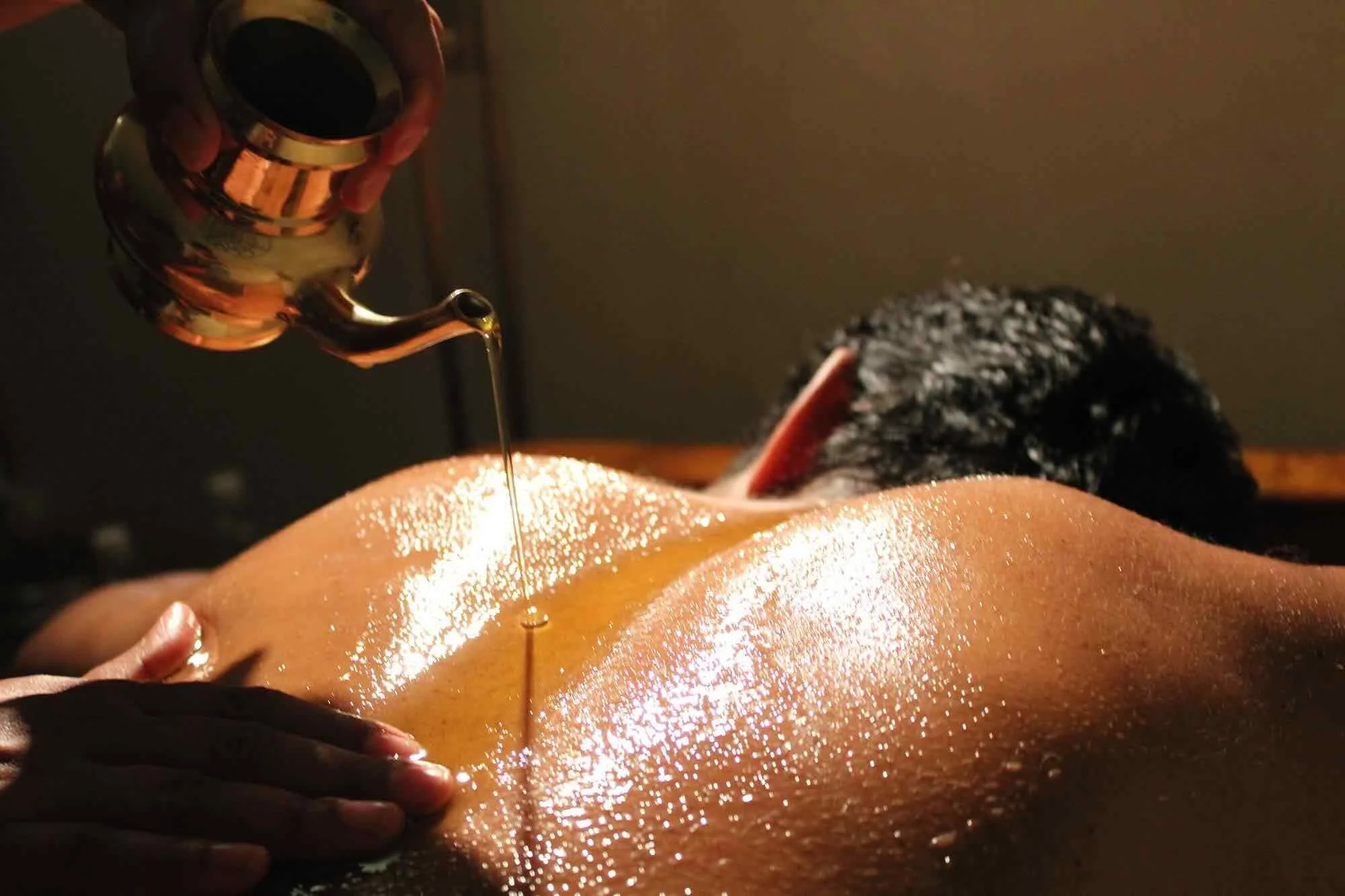 Oil from a brass vessel is poured onto a person's back during massage therapy at Ayurvedic Health Center .