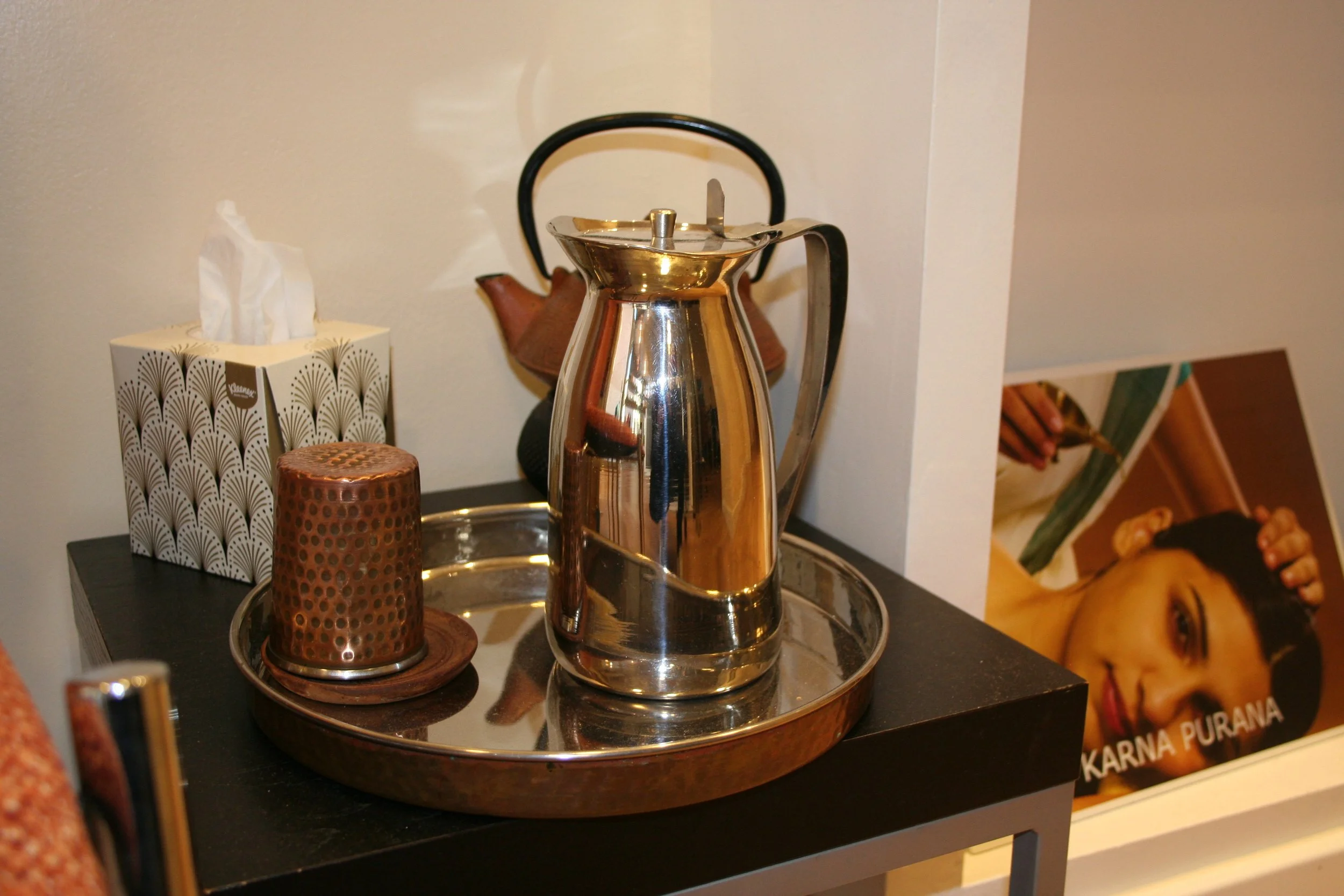 Jarra de acero inoxidable, tetera de cerámica marrón, caja de pañuelos y taza en bandeja de metal en una mesa negra, poster de Karna Purana en el Centro Ayurveda Barcelona