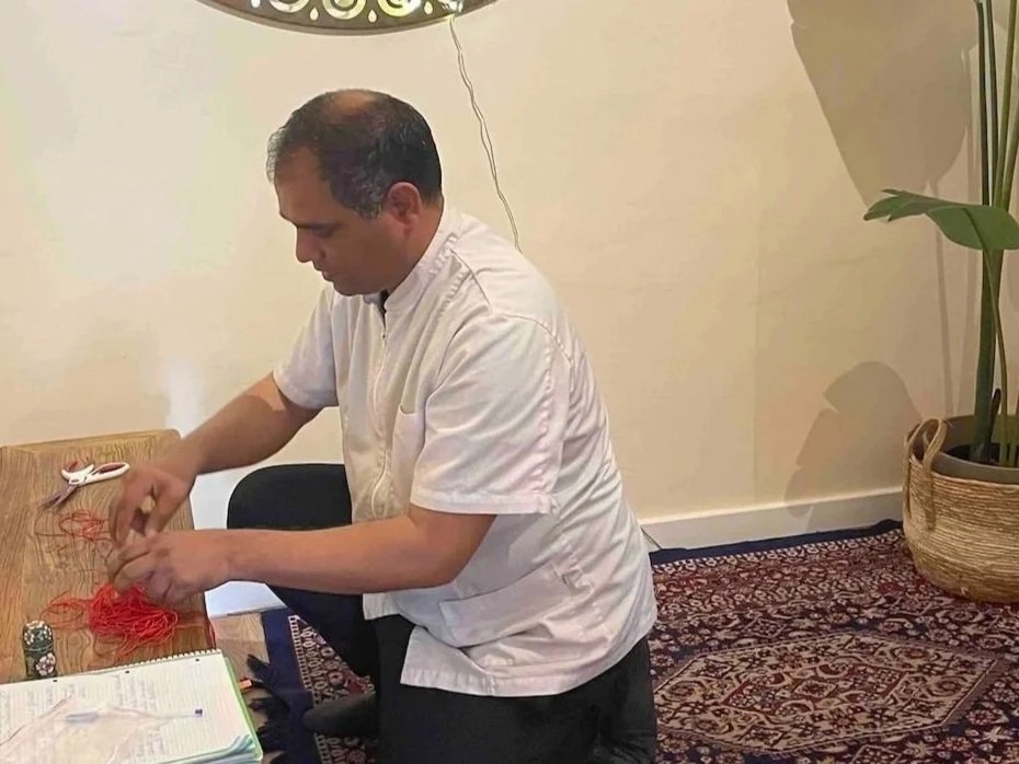 Hombre en una habitación, sentado en una mesa de madera, haciendo manualidades con hilo rojo. Tiene un libro abierto y una pequeña figura de dados en la mesa. Al fondo, una lámpara y una planta en una maceta.