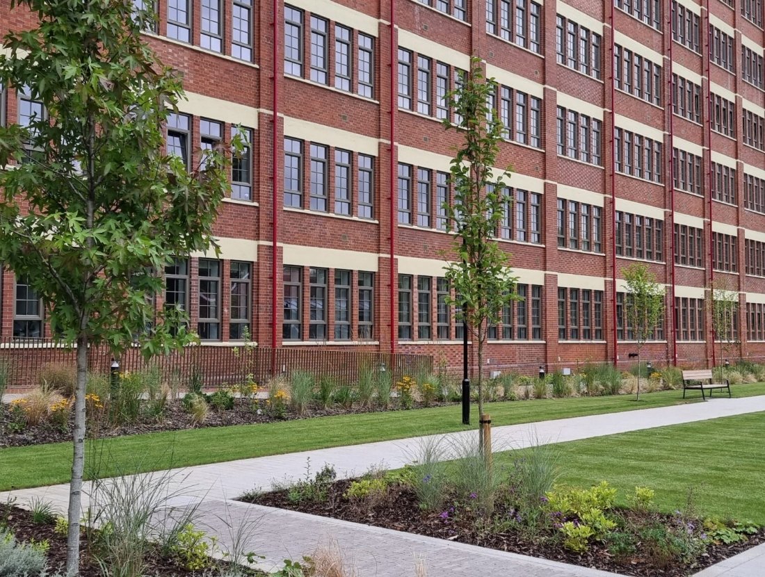 A brick building with multiple windows, surrounded by a landscaped lawn with young trees, shrubs, and benches, and a paved walkway.