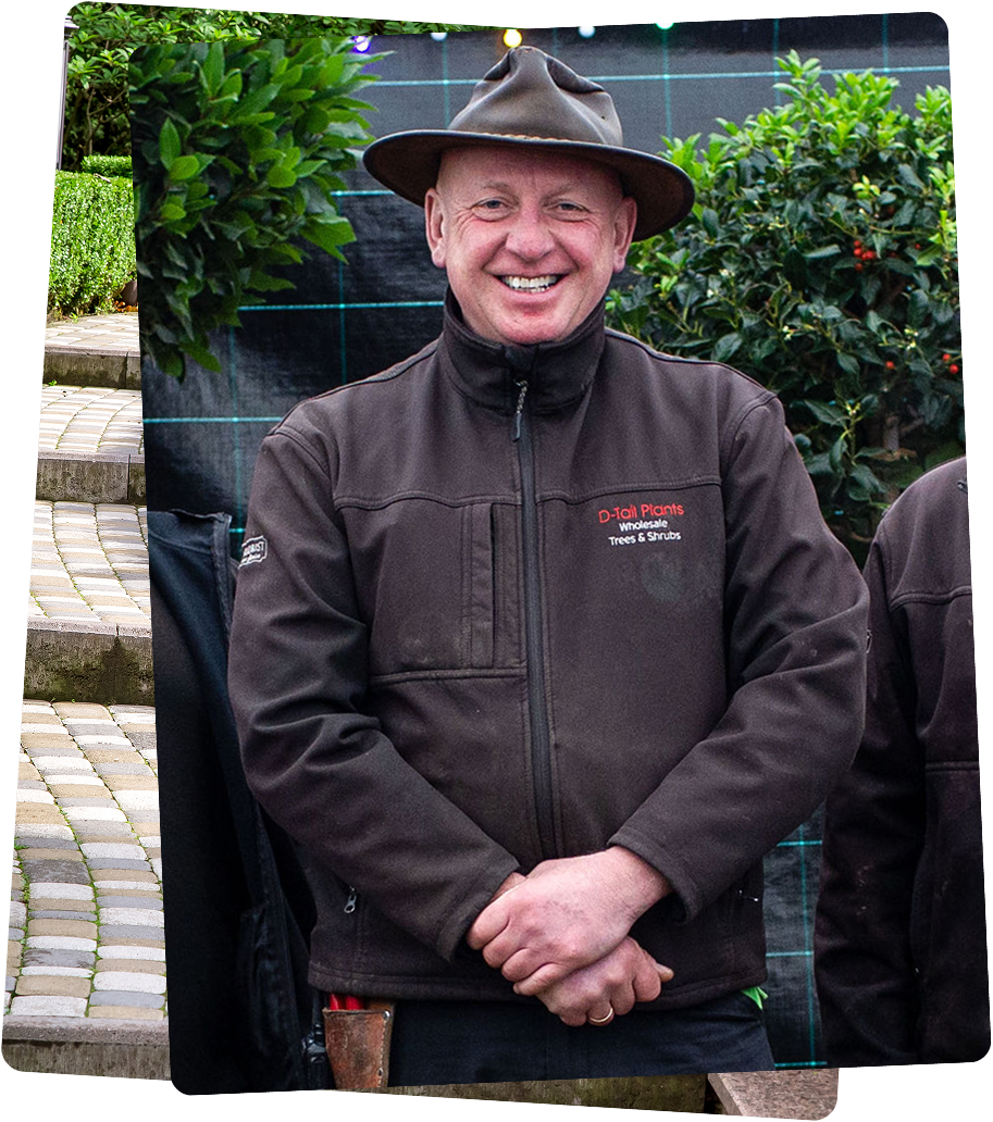 A man smiling outdoors, wearing a brown wide-brim hat and a dark brown jacket with a logo that reads 'D-tall Plants Wholesale Trees & Shrubs' on the chest, with greenery and bushes in the background.