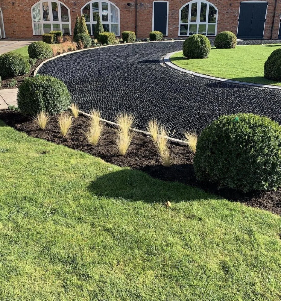 Landscaped front yard with neatly trimmed bushes, a curved garden bed with decorative grass plants, a brick house with large arched windows, and a black gravel driveway.