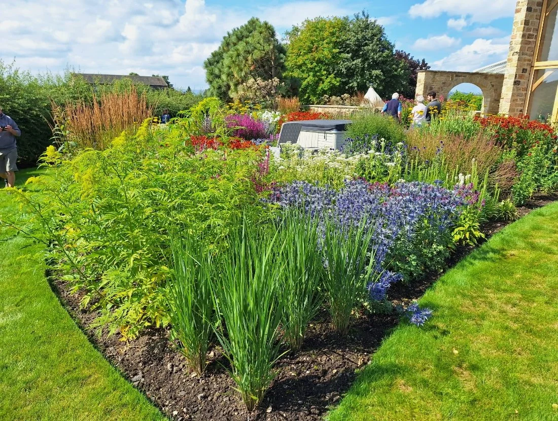 A colorful garden with a variety of blooming flowers and plants, beside a stone building with archways, and people walking and enjoying the outdoor space.