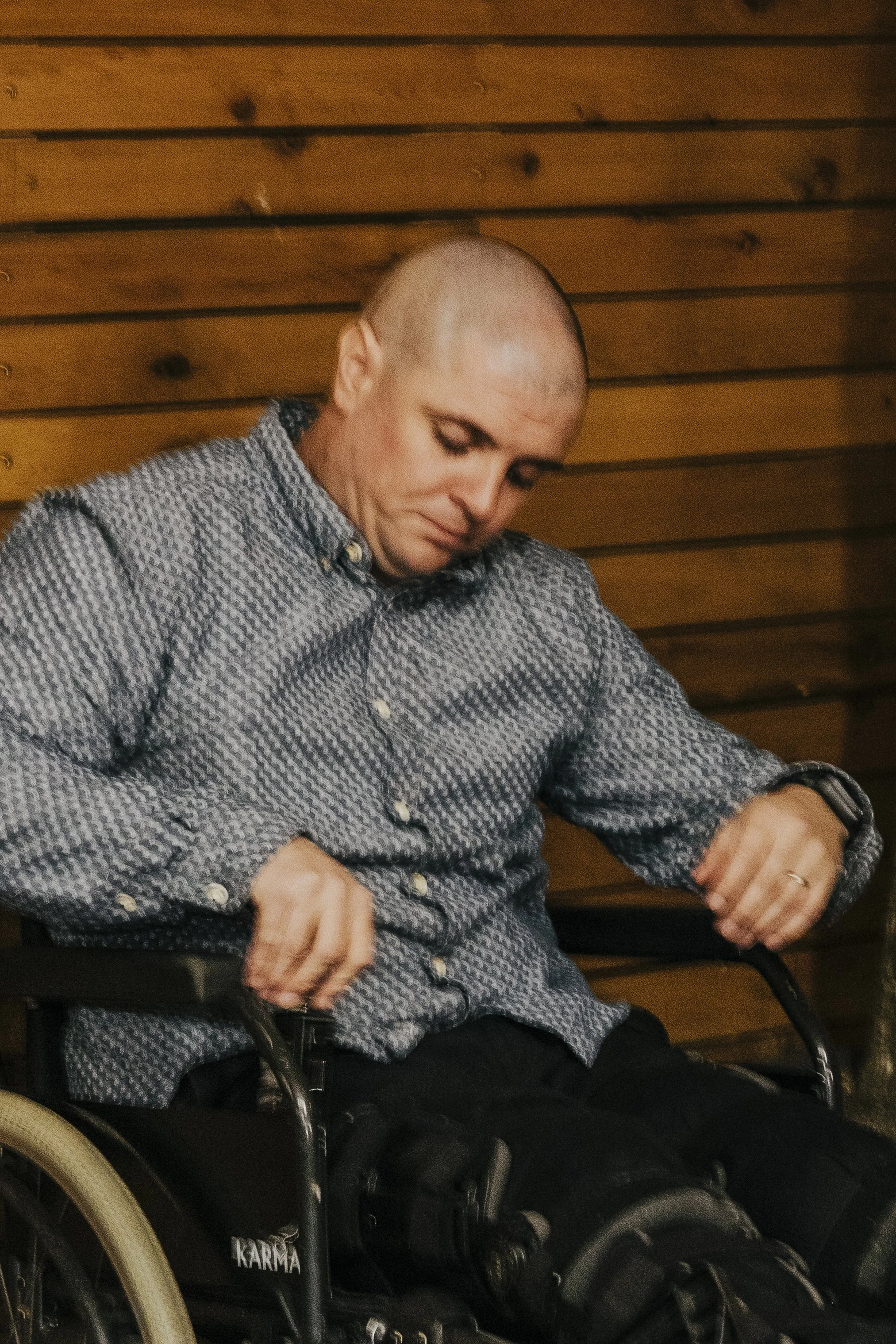 A man with a shaved head sitting in a wheelchair with a wooden wall behind him, wearing a patterned button-up shirt and looking down.