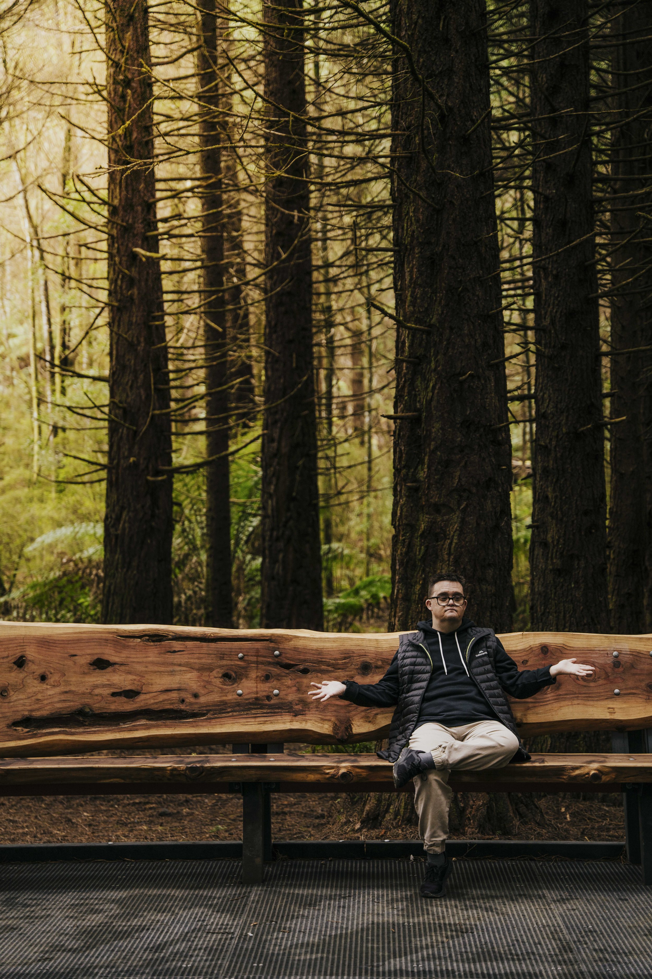 A person sitting cross-legged on a wooden bench with arms outstretched in a forest setting, surrounded by tall trees.