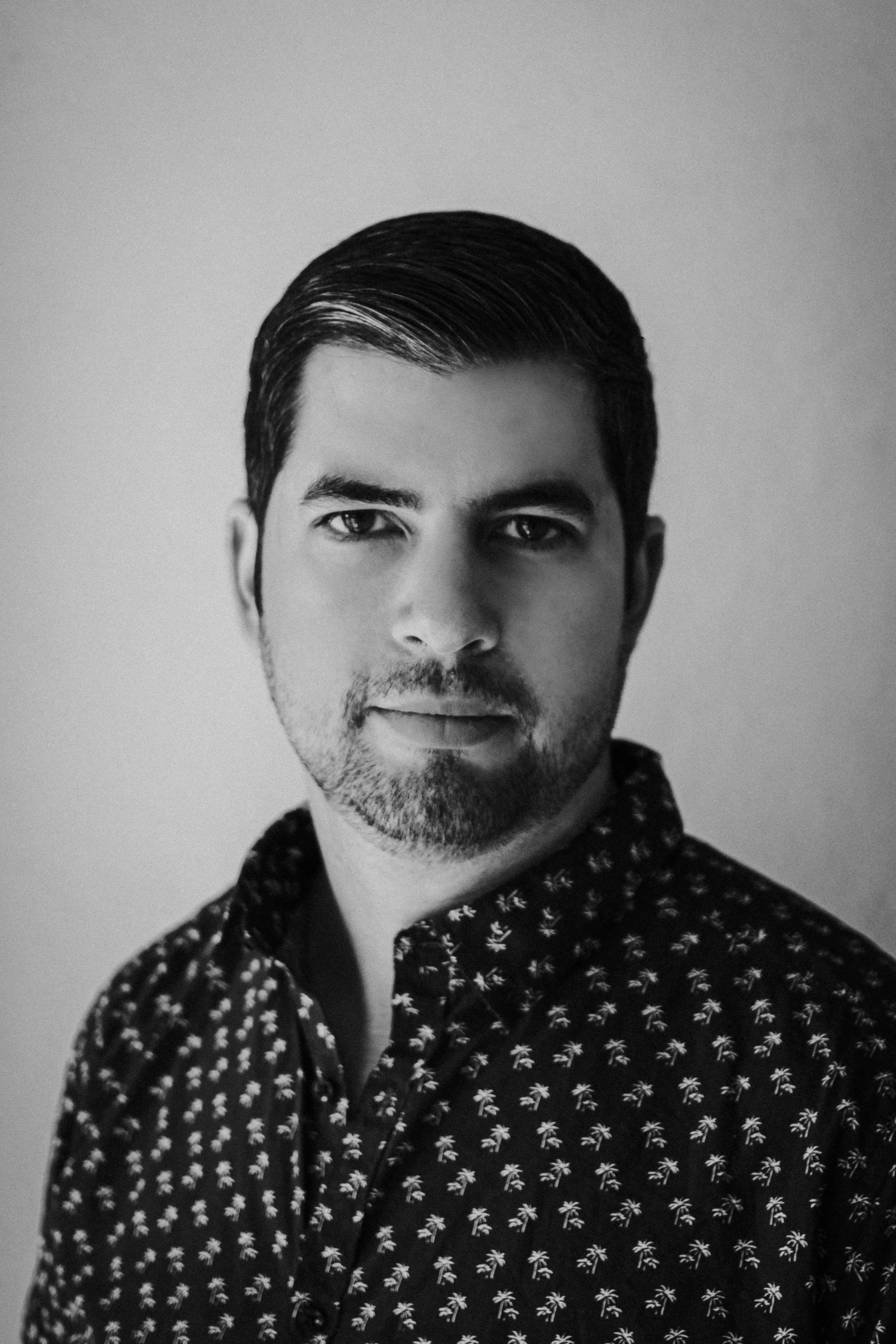 Black and white portrait of a man with dark, combed hair and a trimmed beard, wearing a patterned button-up shirt with small palm tree designs, against a plain background.