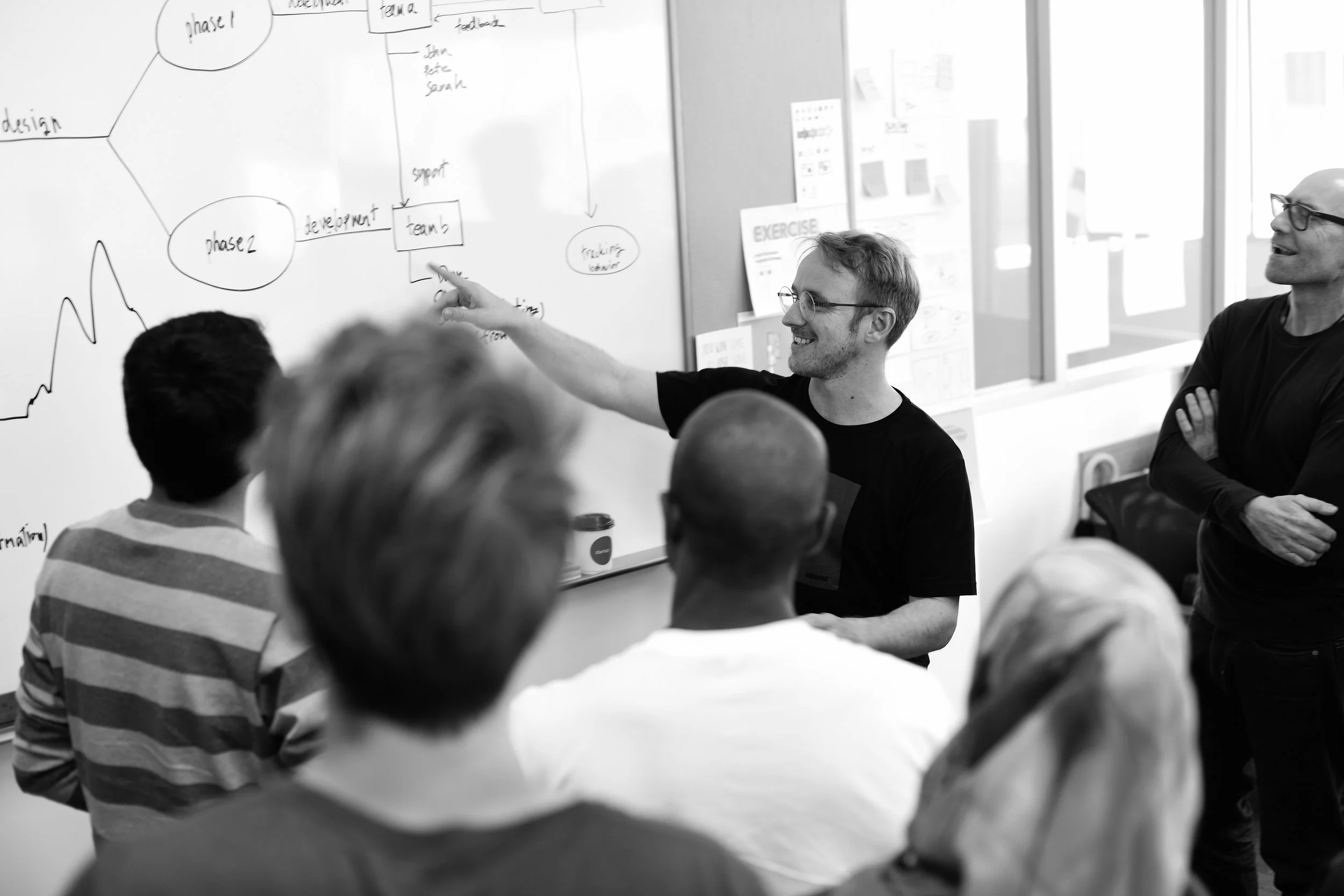 Black and white photo of a man leading a workshop, writing on a whiteboard in front of a group.