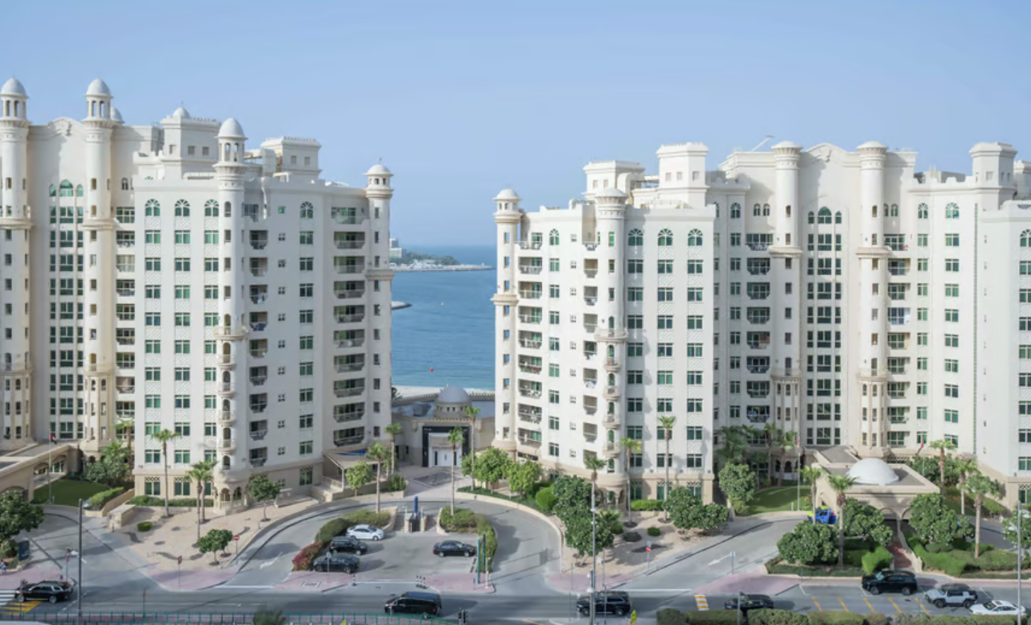 Twin white high-rise buildings near a waterfront with palm trees and parked vehicles in the foreground.
