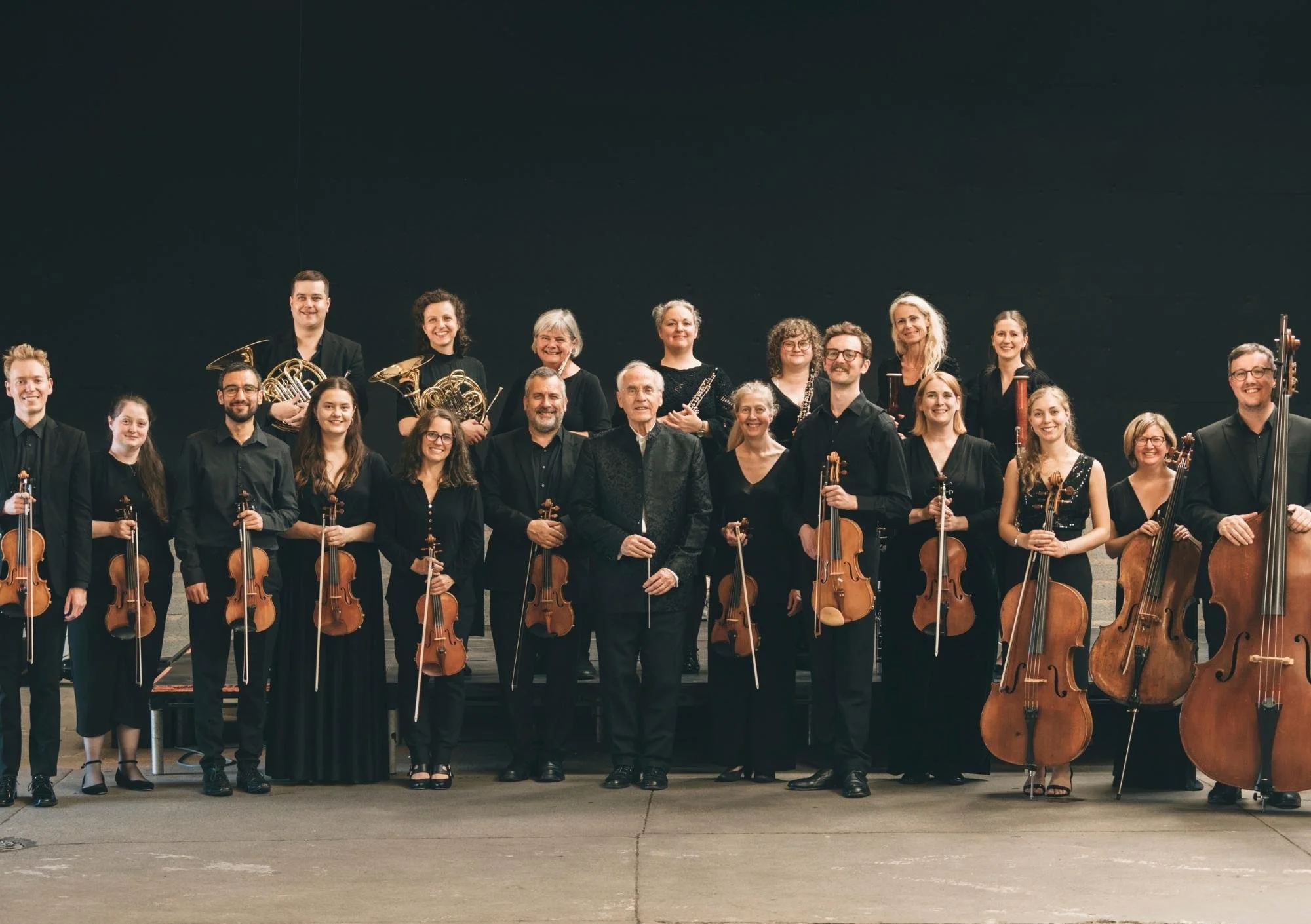 a range of people are standing side by side with some people in the back. they are all wearing black and holding their instruments