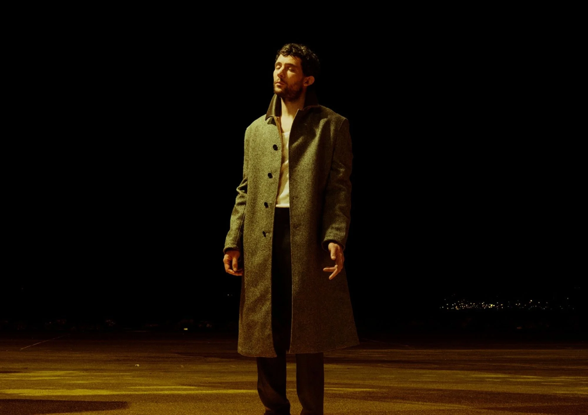 A man stand with his eyes closed in the middle of a dark street and he is wearing a trench coat