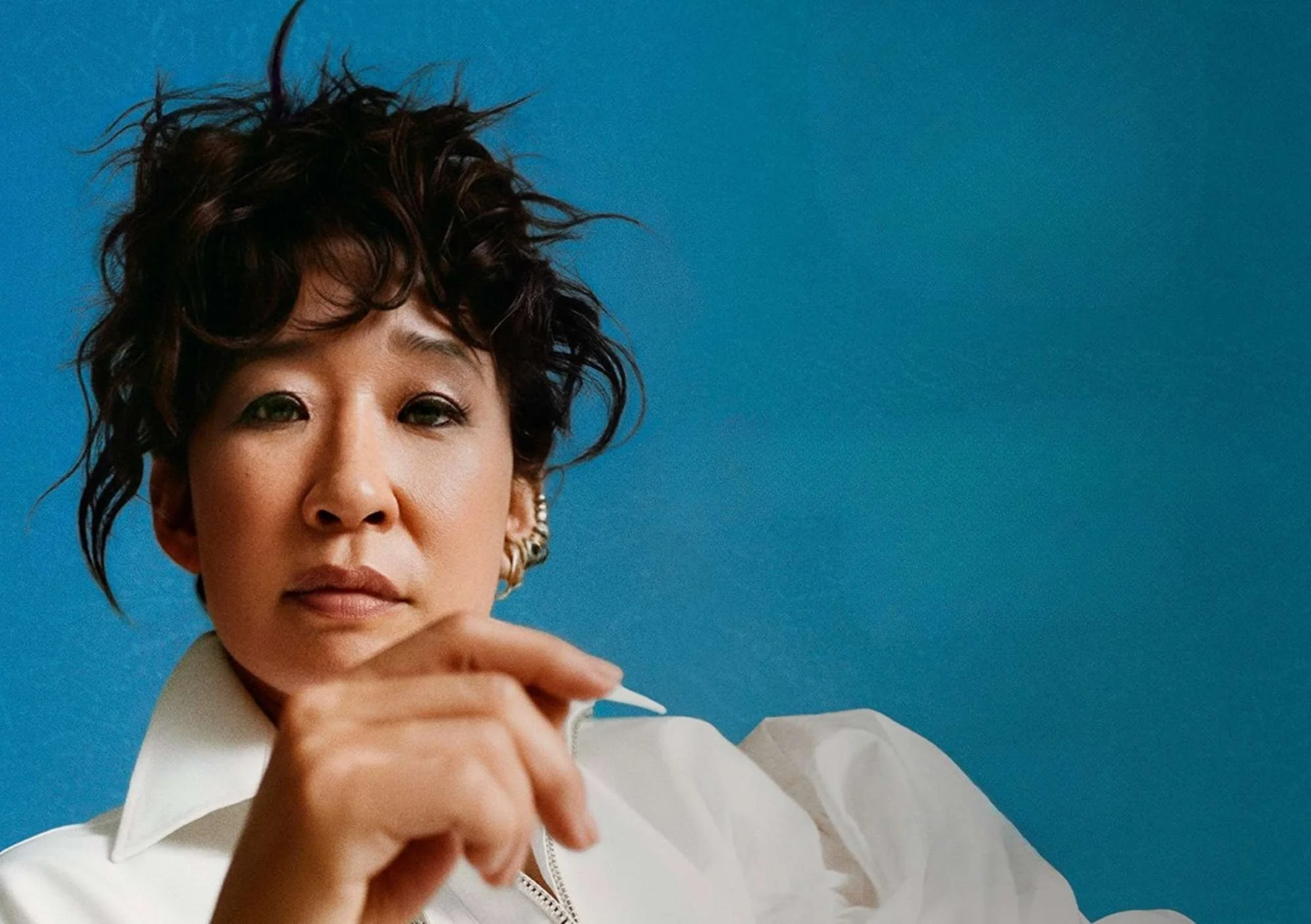 a dark haired women is wearing a white shirt and gold earrings, she is against a blue background