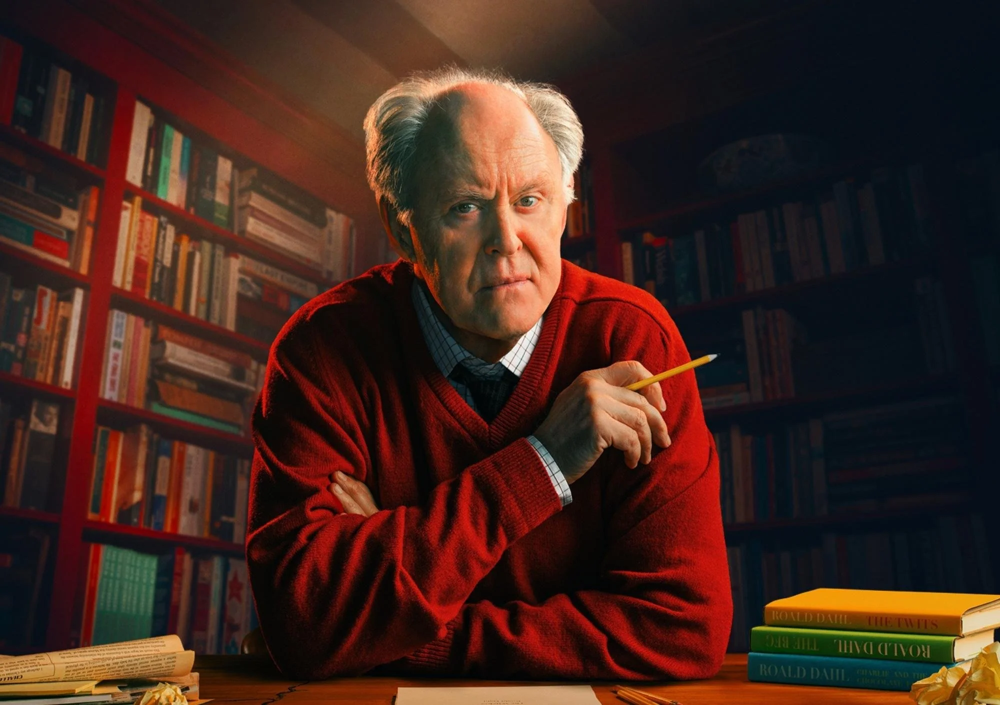 A balding man is wearing a red sweater with a backdrop of bookcases filled with books