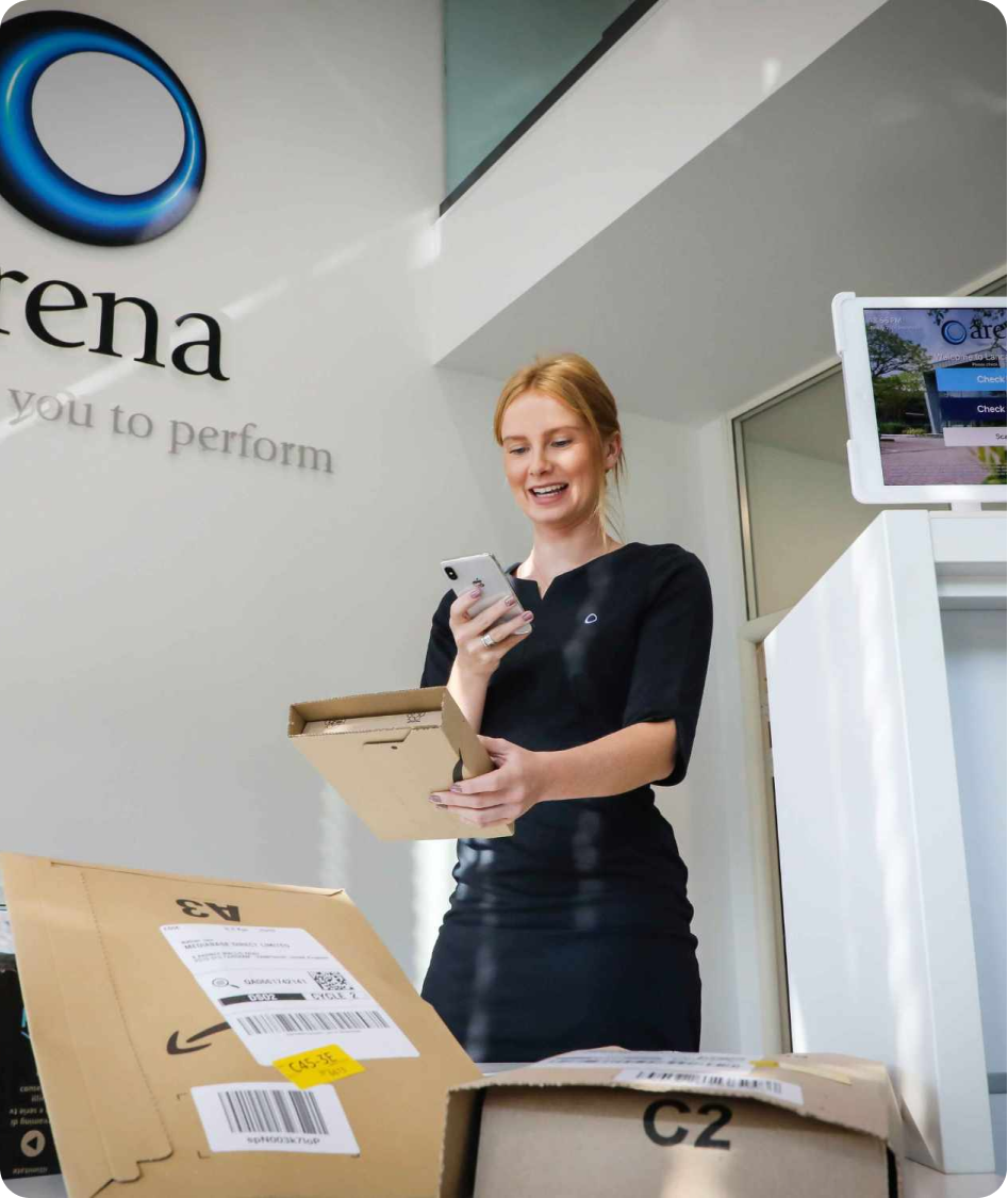 Reception staff member using a smartphone to process a parcel delivery, with visitor management device visible in the reception area.