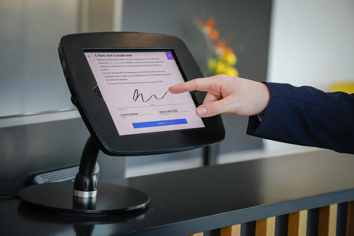 An iPad in a black stand displaying the document signing feature. A finger can be seen coming in from the right to sign the document