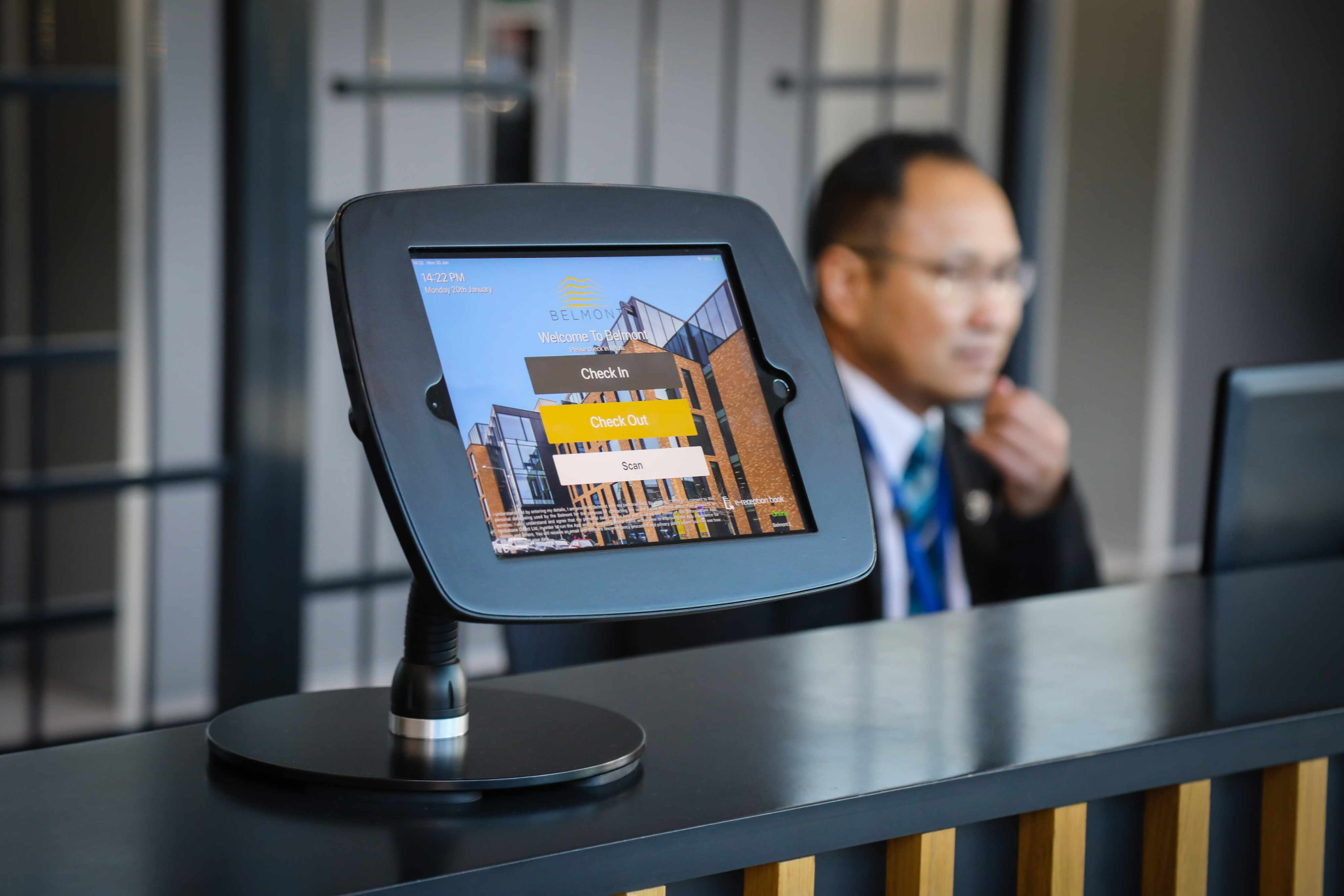Custr check-in system mounted to a reception desk showing visitor check-in, check-out, and scan options, with a staff member working in the background.