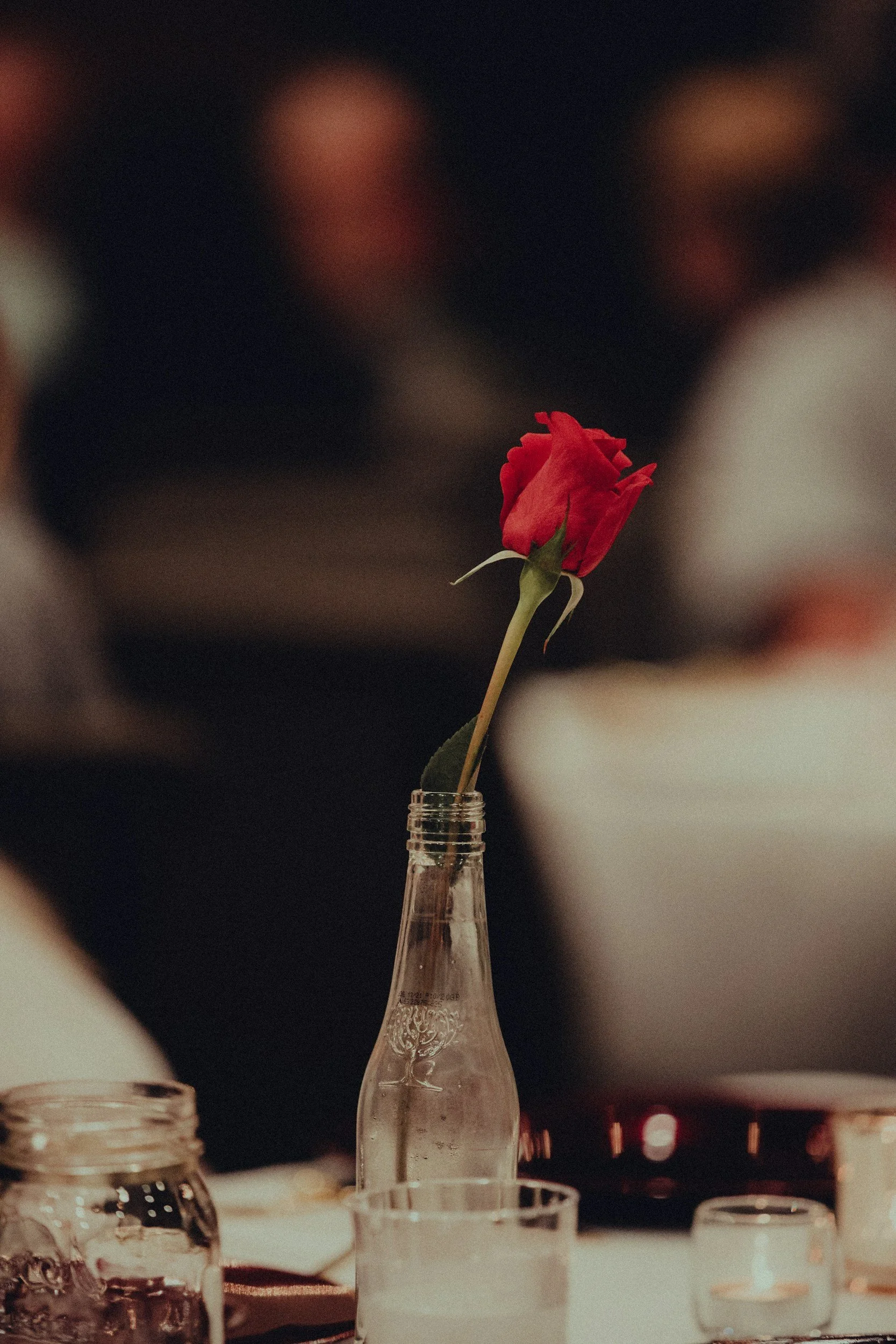 A single red rose in bloom in a glass bottle, with blurred background of people and table setup.