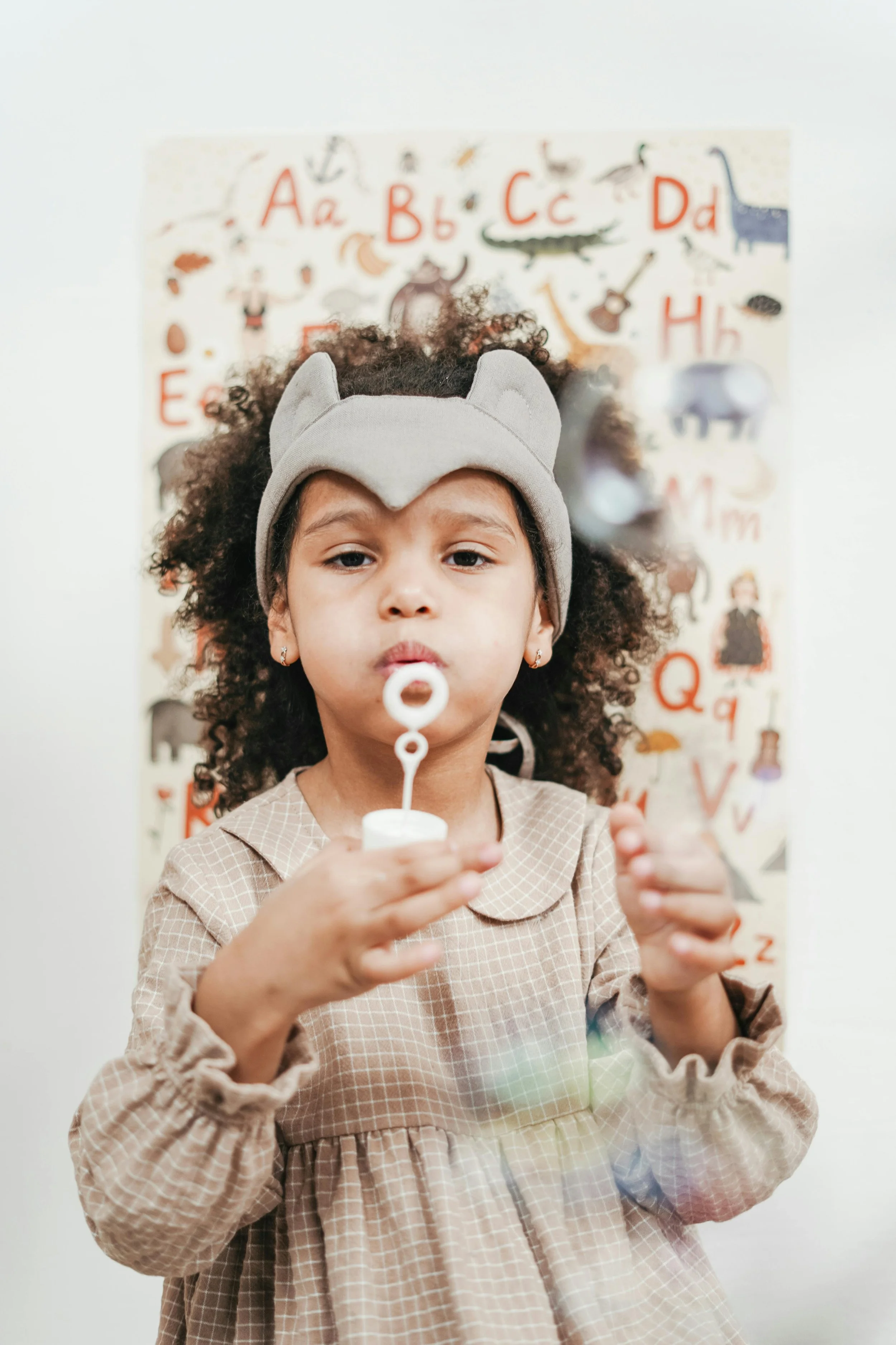 Young girl with curly hair blowing bubbles, wearing a beige checkered dress and a gray cat ear headband, standing in front of an alphabet poster with animal and object illustrations.