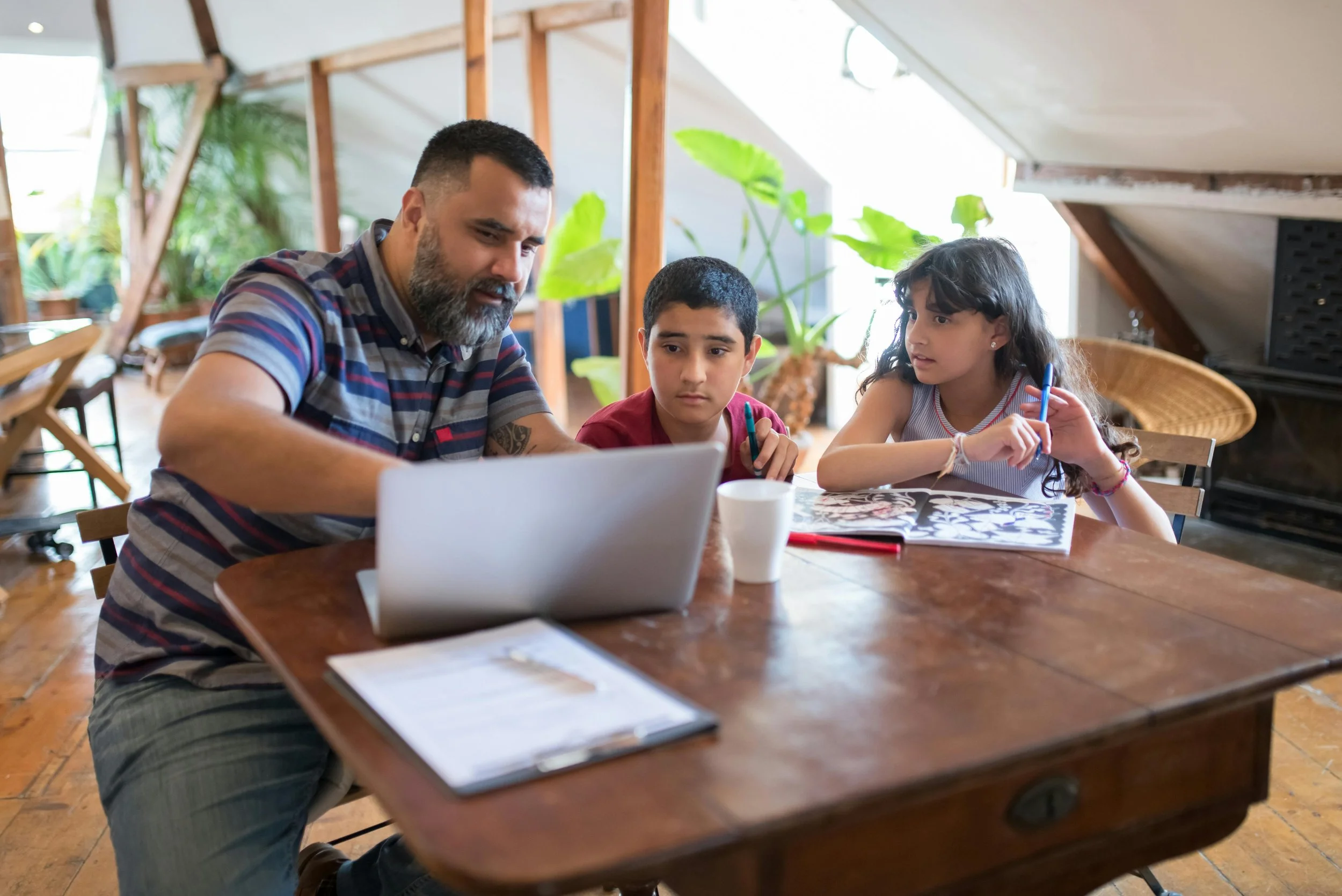 A man with two children sitting at a wooden table with a laptop and notebooks, in a room with plants and wooden decor.