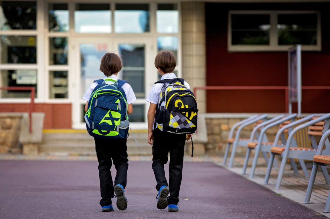 Two young boys wearing school uniforms and backpacks walking toward a school building with large windows and benches outside.
