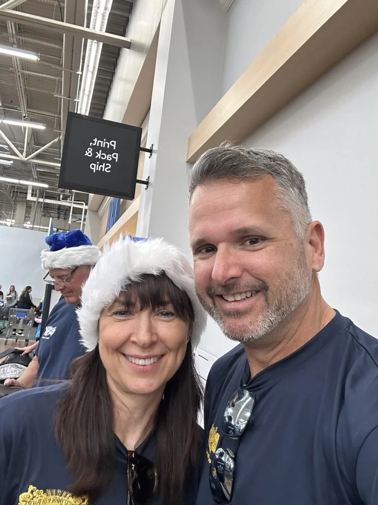 Two smiling adults, a woman wearing a Santa hat and a man, taking a selfie in a store decorated for Christmas. They are dressed in navy shirts, and the woman has glasses hanging from her neck. A man in the background is wearing a Santa hat and appears to be working near shopping carts.