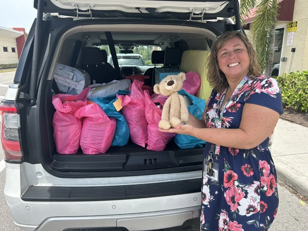 A woman in a floral dress holding a teddy bear, standing in front of an open SUV filled with pink and blue gift bags.