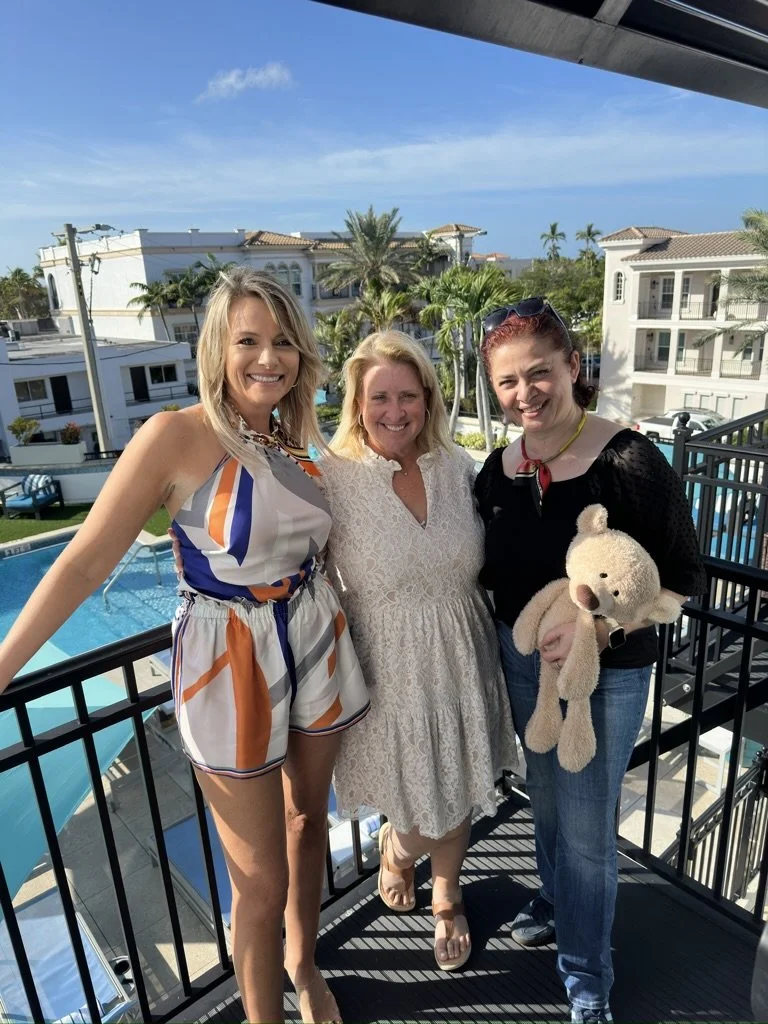 Three women smiling on a balcony overlooking a pool and palm trees at a sunny day in a residential area.