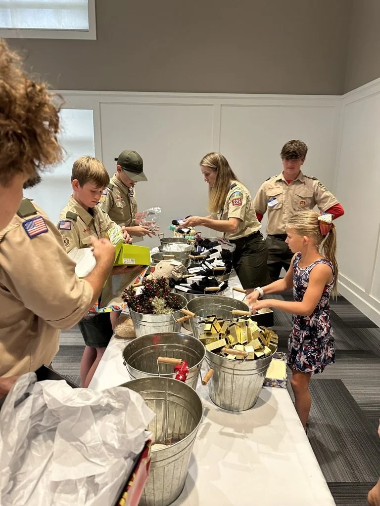 A group of both young girls and boys, some in scout uniforms, standing at a table filled with various items, including buckets of soap, soap molds, and cleaning supplies, preparing for an activity or event.