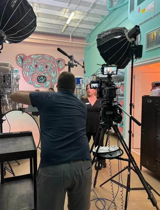 A woman being recorded by a camera and lighting equipment in a colorful room with a large bear face mural on the wall.