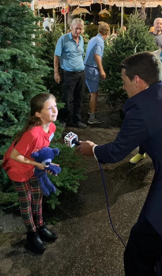A girl being interviewed by a news reporter at a Christmas tree lot, holding a blue stuffed toy, with several people and Christmas trees in the background.