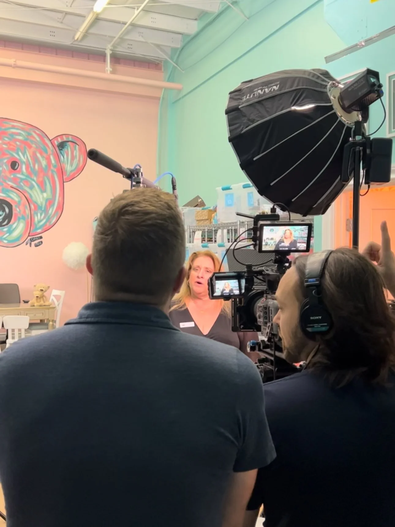 A woman being interviewed or recorded with a professional camera and lighting equipment, in a room with colorful walls and a bear mural.