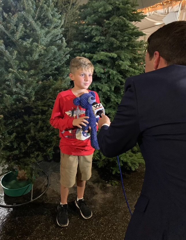A young boy in red shirt and khaki shorts being interviewed with a microphone held by a person in a dark suit in front of Christmas trees.
