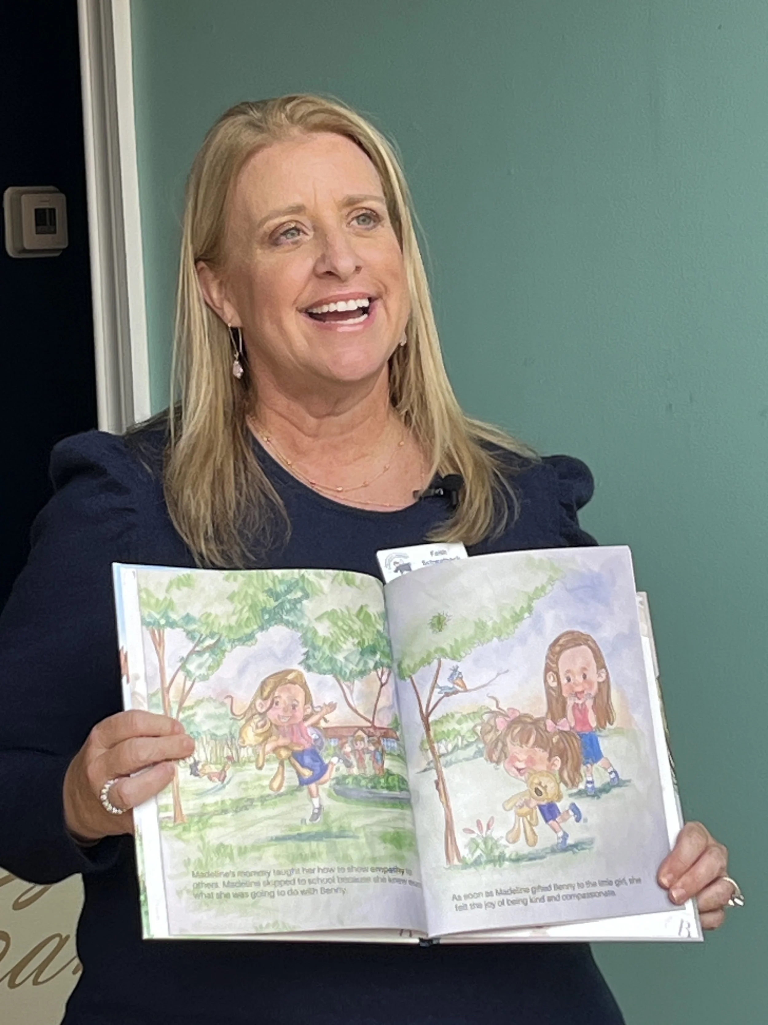 A woman with blonde hair holding up an illustrated children's book open to a page showing children playing outside under trees, smiling.