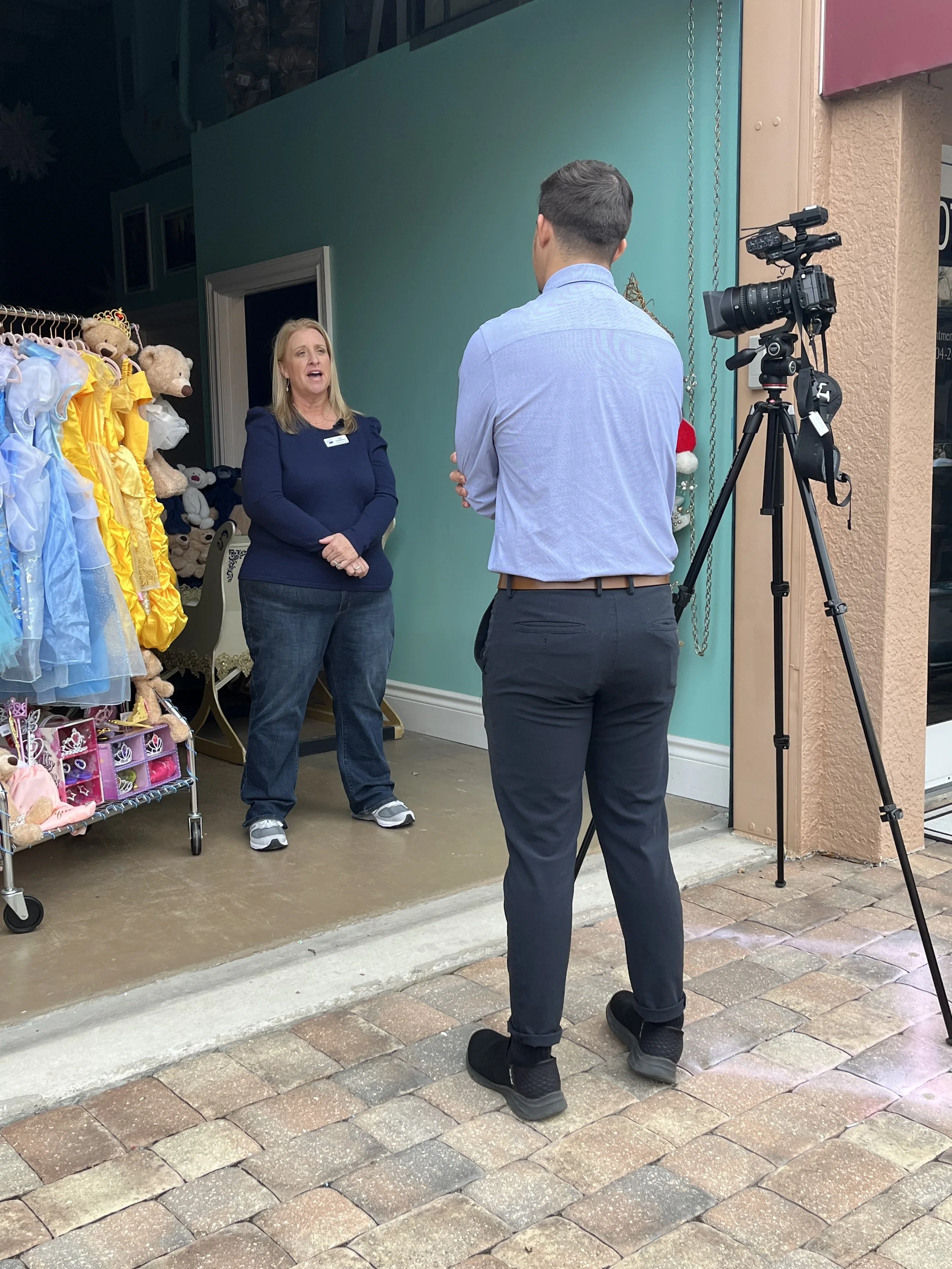 A woman being interviewed on camera outside a storefront with toys and costumes on display.
