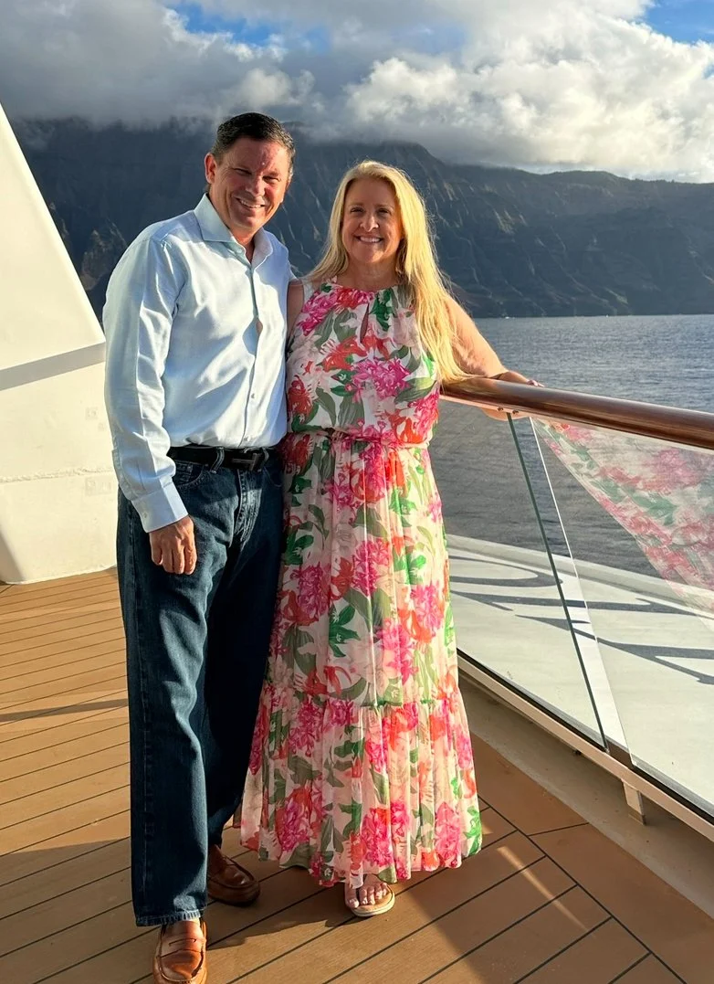 A smiling man and woman standing together on a ship's deck with a scenic view of mountains and water in the background. The man is wearing a white shirt and jeans, and the woman is in a colorful floral dress.