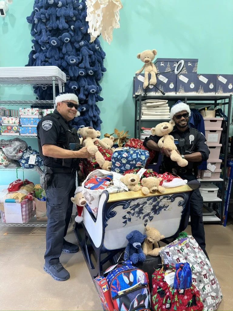 Two police officers wearing Santa hats and sunglasses standing in a store surrounded by teddy bears, gift bags, and holiday decorations during Christmas.