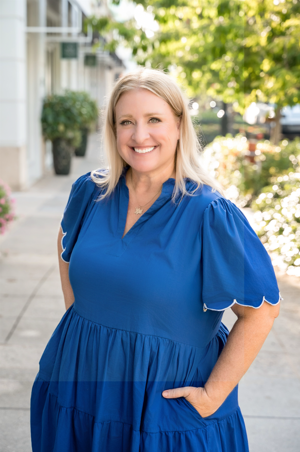 A smiling woman with blonde hair wearing a blue dress, standing outdoors on a sidewalk with trees and buildings in the background.