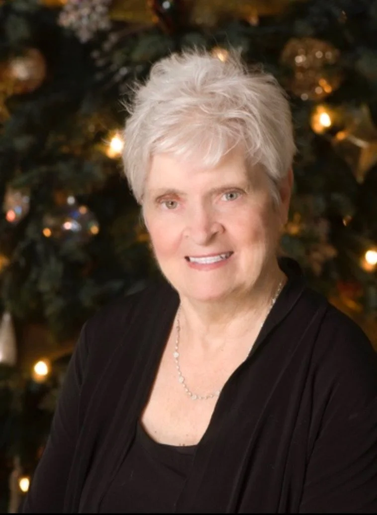 Beverly with short, silver hair smiling, wearing a black top and necklace, standing in front of a decorated Christmas tree with ornaments and lights.
