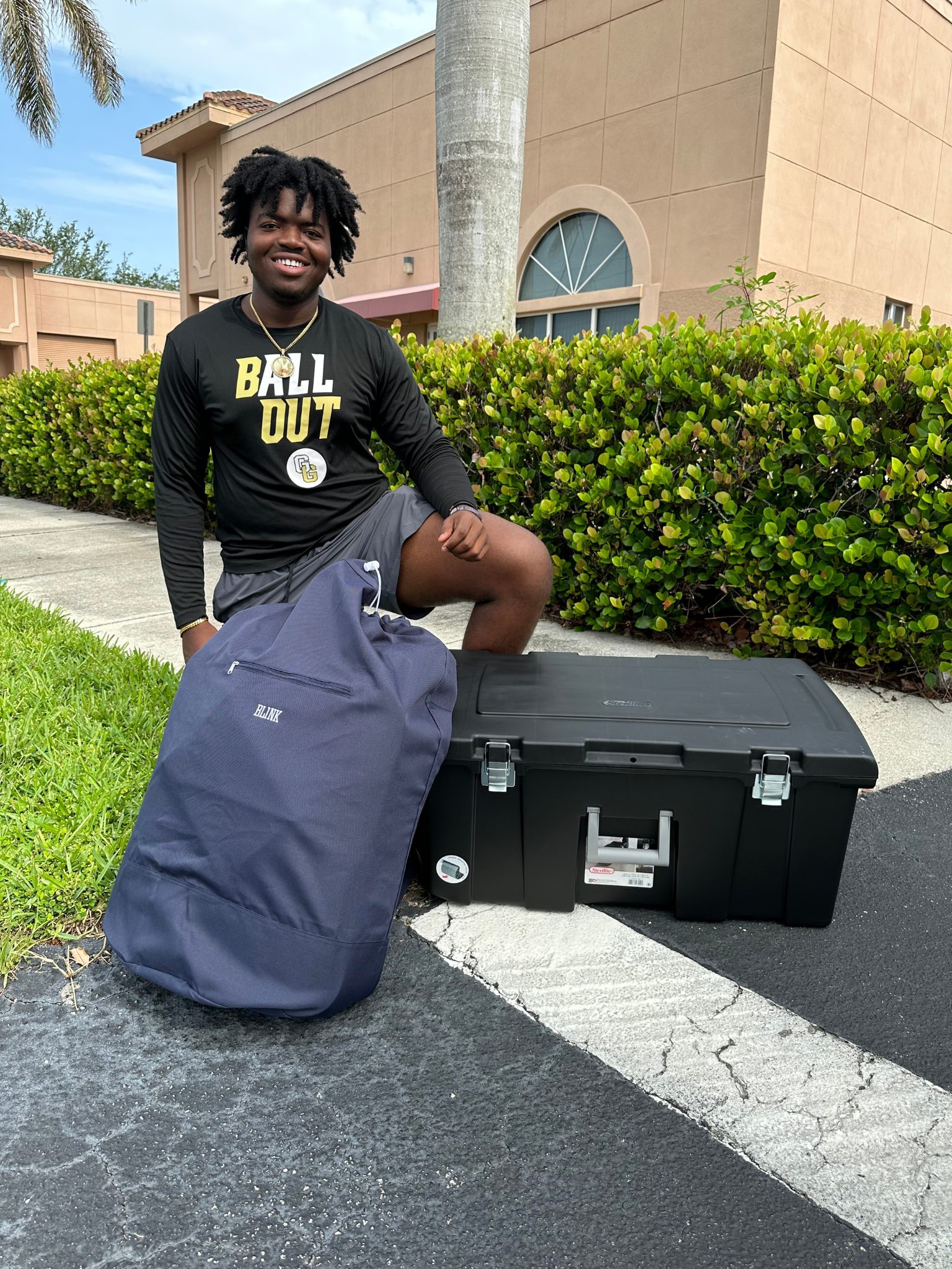 A young man kneeling outdoors next to a large black equipment case and a navy backpack, smiling at the camera, with a beige building, palm trees, and green bushes in the background.