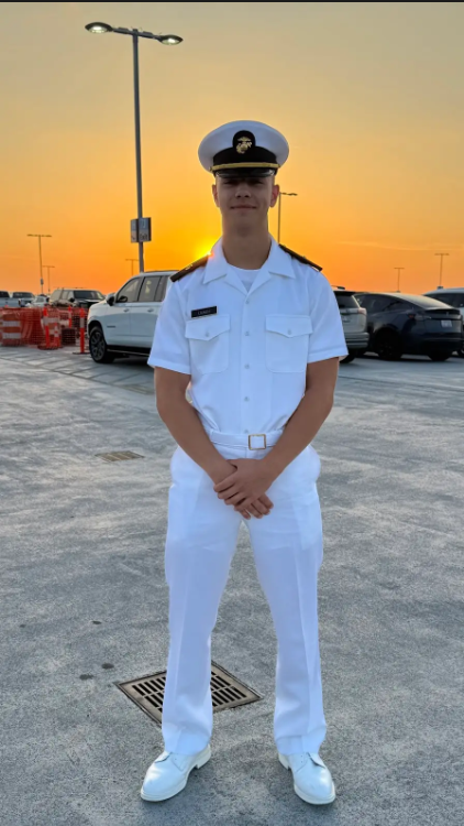 Young man in a white naval uniform standing in a parking lot during sunset.