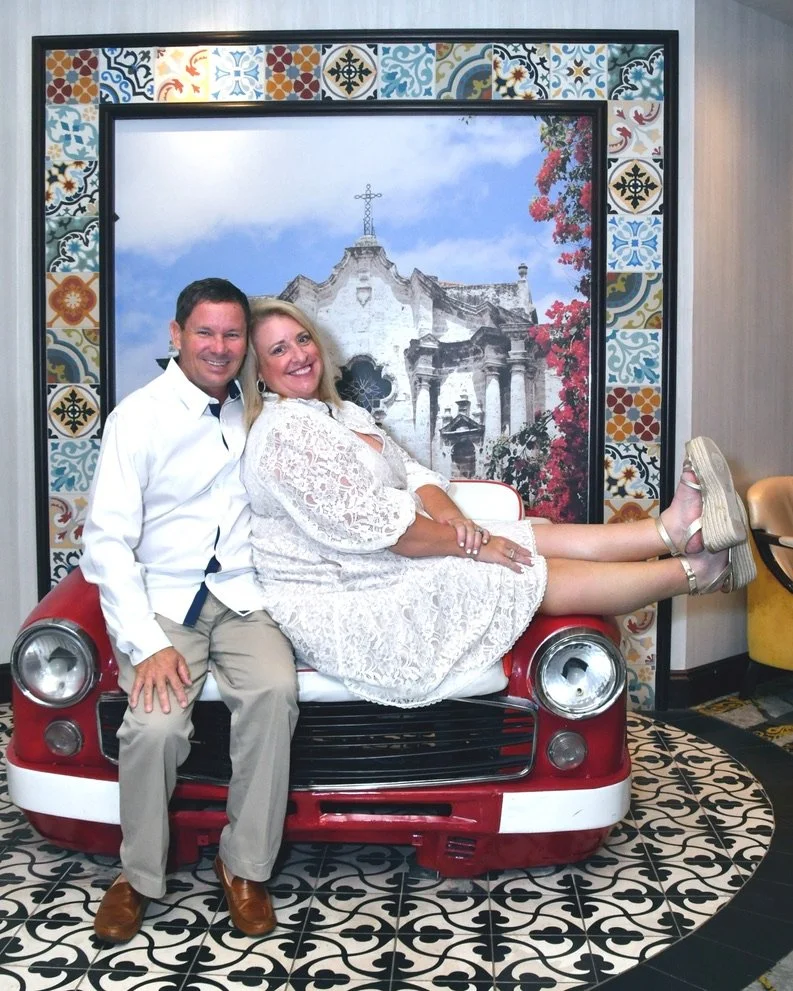 A smiling man and woman sitting on a red car-shaped bench, with the woman lying across their laps and posing with her legs outstretched. The background features a decorative framed image of a church with a cross and colorful patterned tiles.