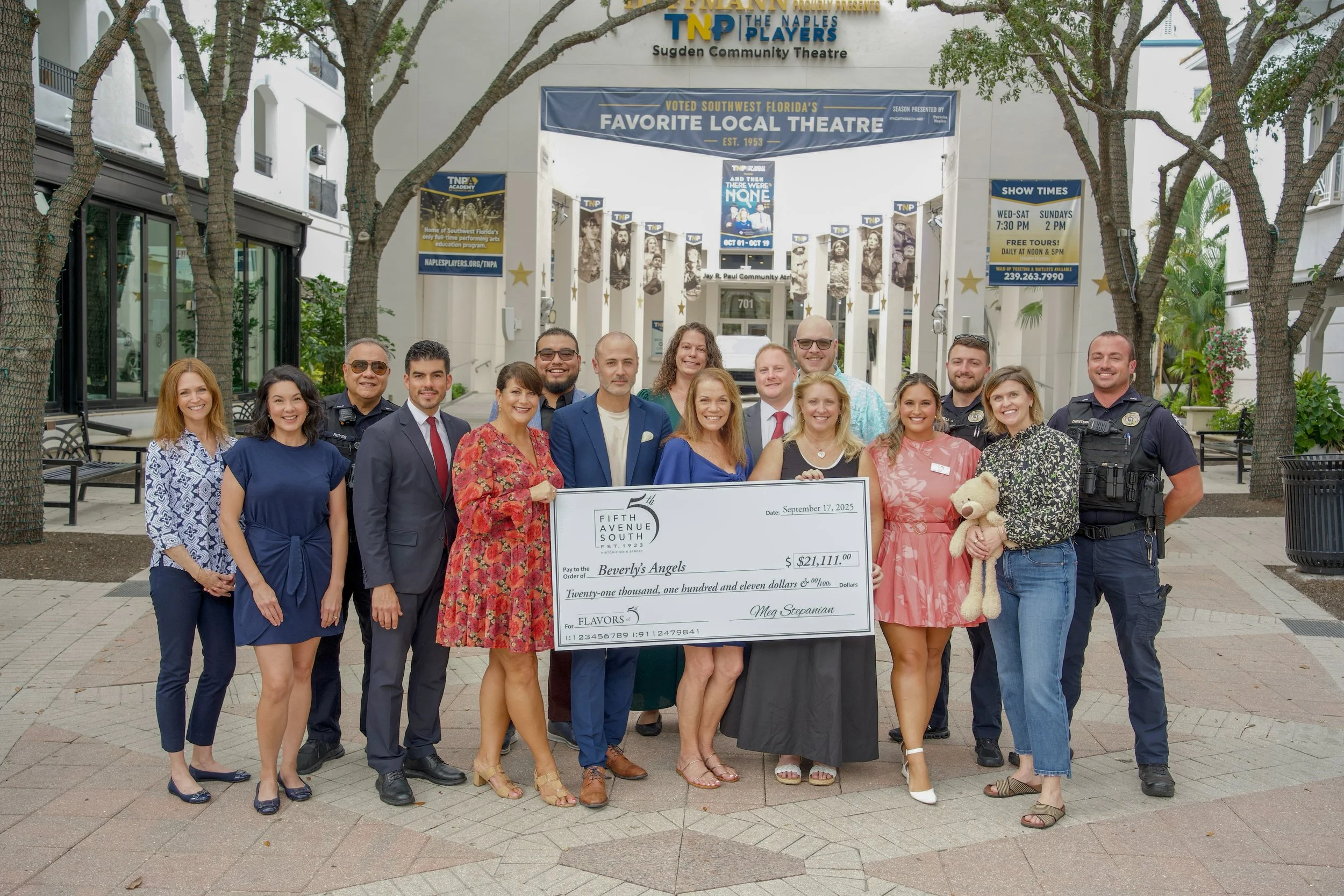 Group of people standing outdoors holding a large check in front of a theater entrance with banners and trees.