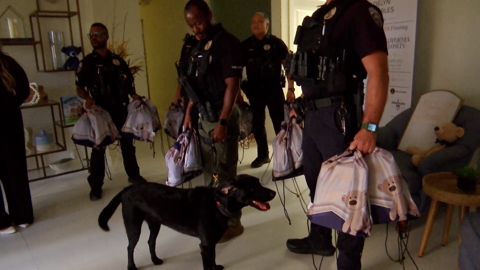 Four police officers standing indoors with a black police dog, all carrying bags, with a beige sofa and a teddy bear in the background.