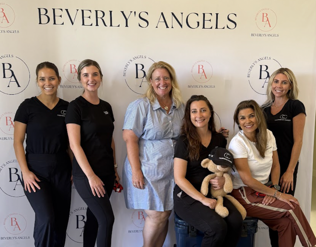 Group of six women, some in medical scrubs, smiling and posing for a photo in front of a Beverly's Angels banner. One woman is holding a teddy bear.