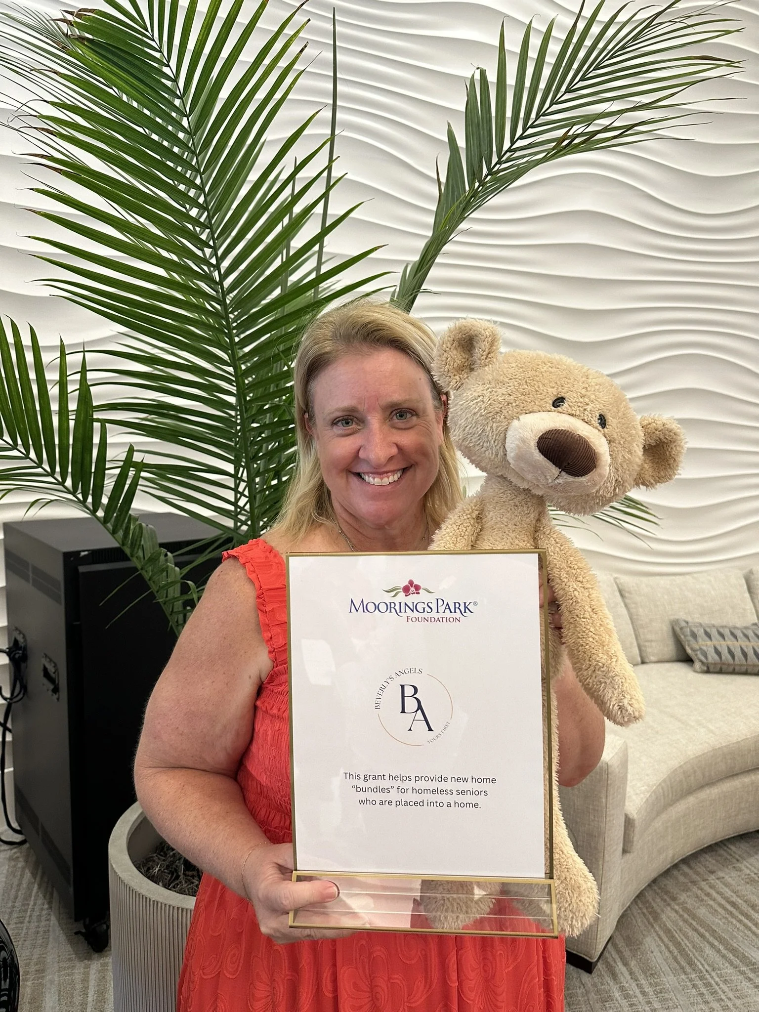 A smiling woman in a red dress holding a framed certificate and a teddy bear, standing in front of a large green plant and textured white wall.