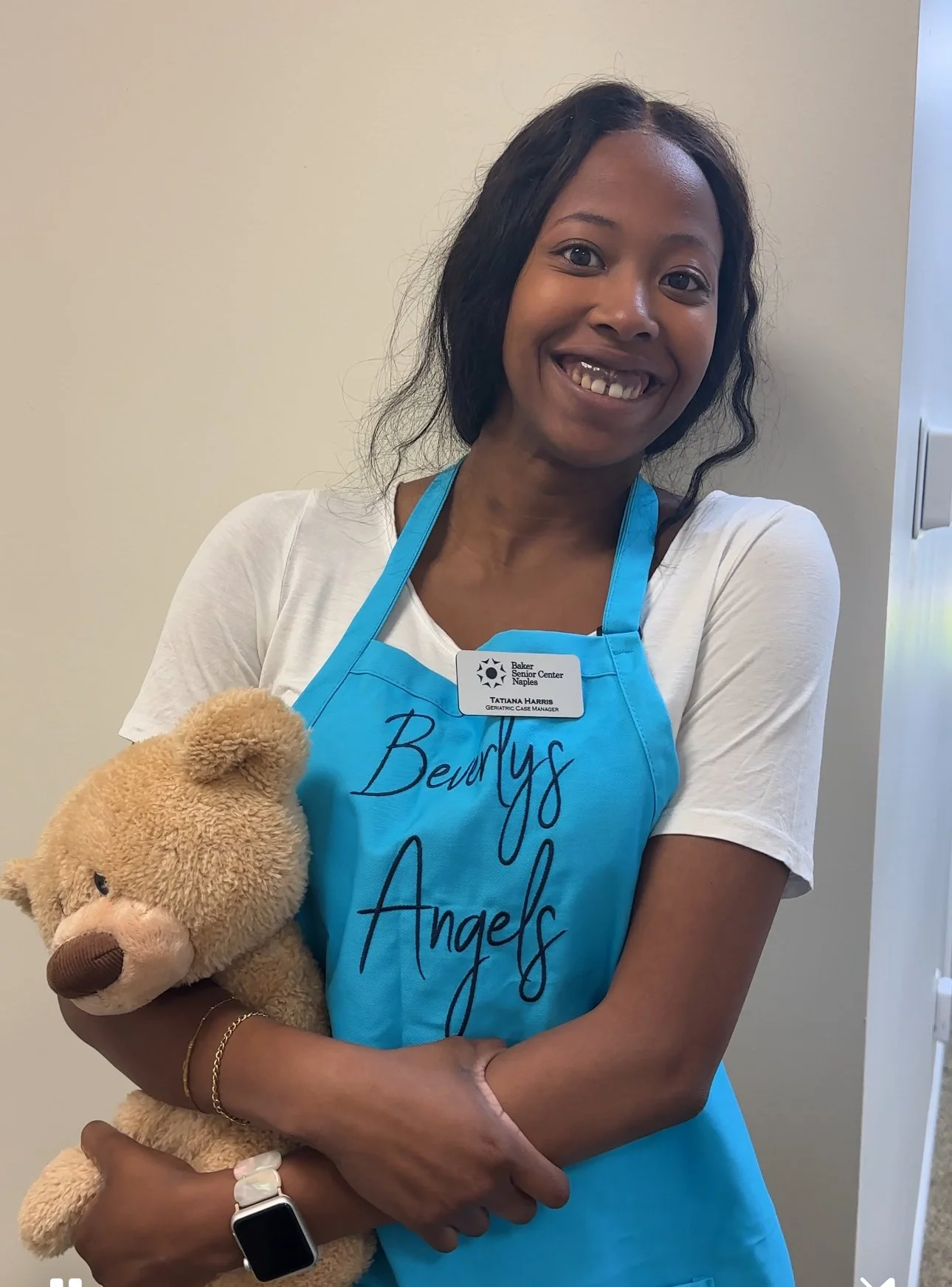 A smiling woman with dark hair, wearing a white t-shirt and a blue apron with the text "Beaulys Angels," holding a teddy bear, standing indoors against a beige wall.